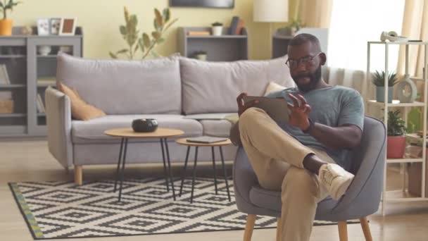 アフリカ系アメリカ人のフリーランサーの肖像画 リビングルームのアームチェアに座りながらデジタルタブレットを使用 — ストック動画