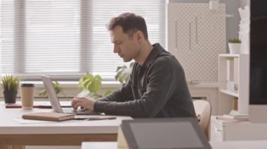 Bilgisayarla çalışan beyaz erkek girişimcinin ofis masasında oturmasının yan görüntüsü yavaşlıyor