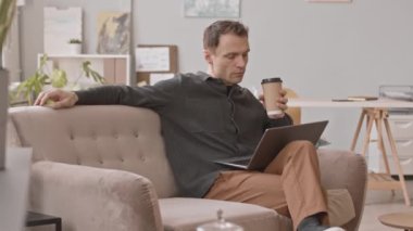 Diz üstü bilgisayarda çalışan ve modern ofiste kadife bej koltukta otururken kahve fincanından kahve yudumlayan erkek girişimci.