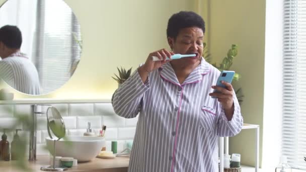 Black Woman Striped Pajamas Scrolling Smartphone While Brushing Her Teeth — Stock Video