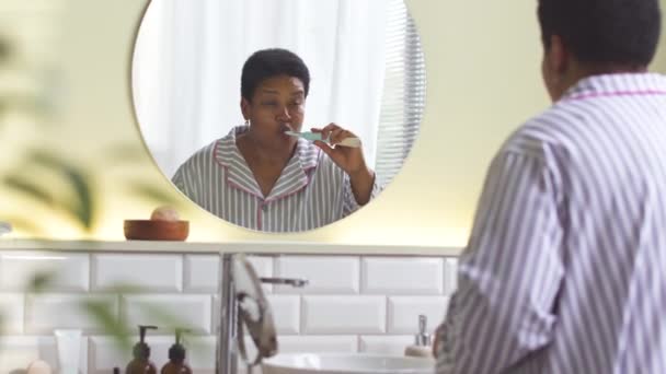 身穿条纹睡衣的中年黑人妇女站在浴室圆形镜子前刷牙 — 图库视频影像