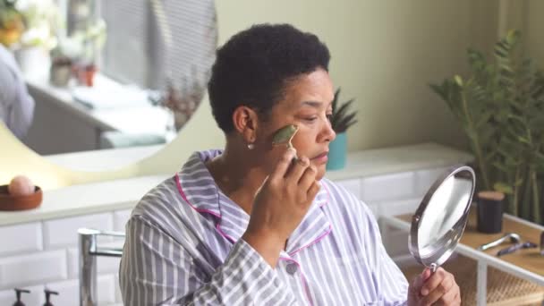 非洲裔美国妇女早上站在浴室里用石英滚筒做面部按摩 并在镜子中看到自己的倒影 — 图库视频影像