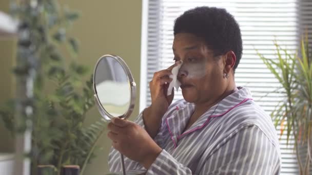 穿着条纹睡衣的中年非洲裔美国妇女一边看着镜子 一边从浴室的脸上摘下黏土面具 — 图库视频影像