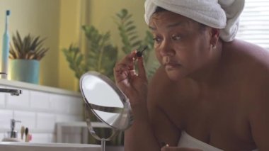 Olgun Afro-Amerikalı kadın sabah makyajını yaparken aynaya bakıyor, rimel sürüyor.