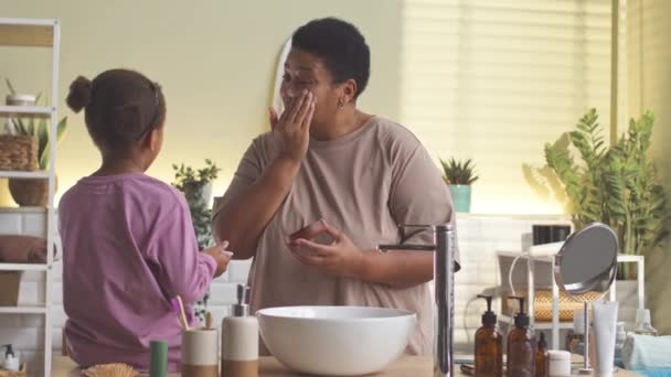 非洲裔美国人成熟的女人和她的小女儿一起在浴室里做日常皮肤护理 在脸上涂奶油 — 图库视频影像