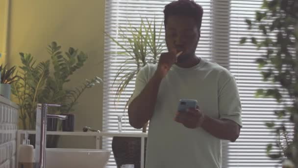 Joven Hombre Afroamericano Navegando Teléfono Inteligente Mientras Cepilla Los Dientes — Vídeo de stock