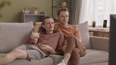 Romantik, genç, beyaz, eşcinsel çift oturma odasında koltukta rahatça oturur televizyon izler ve sohbet ederler.