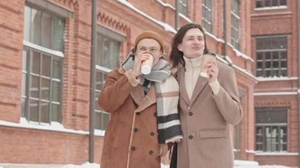 在寒冷的冬日里 两个年轻快乐的男朋友一边在户外散步一边聊天 一边用外卖的咖啡杯喝咖啡 — 图库视频影像