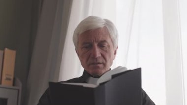 Yetişkin, gri saçlı, papaz tasmalı bir rahibin evde dua okuması.