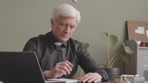 高级灰发牧师身穿黑色衬衫 头领紧盯着摄像机 双手合锁坐在办公桌前的画像 — 图库视频影像