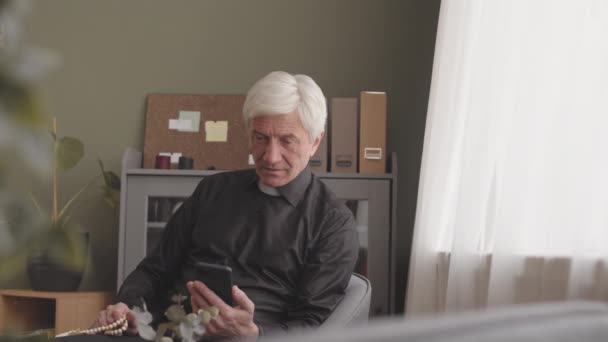 高级灰发牧师身穿黑色衬衫 在智能手机上与教区居民在线视频交谈 在家工作 — 图库视频影像