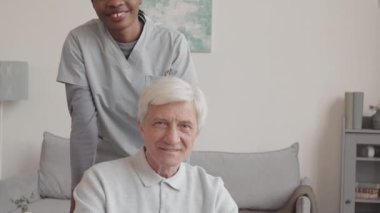 Üniformalı gülümseyen Afrikalı Amerikalı kadın hemşire ve içeride kameraya poz veren beyaz tekerlekli sandalyedeki yaşlı adamın yavaş yavaş portresini indirin.