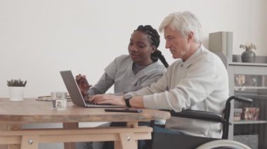 Genç Afrikalı Amerikalı kadın sosyal hizmetler görevlisi, tekerlekli sandalyedeki yaşlı adama dizüstü bilgisayar kullanmayı öğretiyor, rahat adamların evinde masa başında oturuyor.