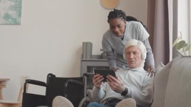 Kablosuz kulaklıklı beyaz bir adam dairesinde oturmuş dijital tablet kullanmayı öğrenirken Afro-Amerikan kadın sosyal hizmet görevlisi ona yardım ediyor.