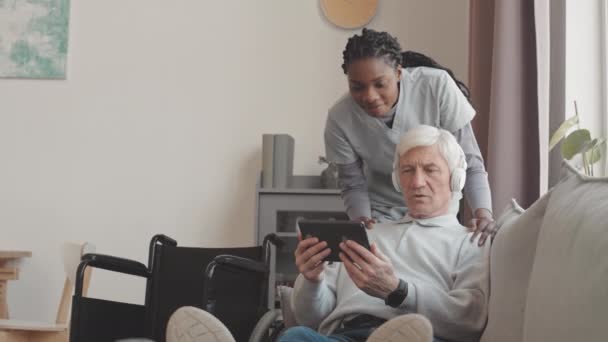 在非洲裔美国女性社会工作者的帮助下 坐在公寓沙发上学习使用数码平板电脑的白种人老年男子在无线耳机中学习使用数字平板电脑 — 图库视频影像