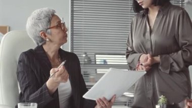 Çizgili ceketli ve gözlüklü olgun bir Asyalı iş kadını proje fikirleri ve şirket sonuçlarını tartışmak için takım toplantısını yönetirken ofis masasında oturuyor.