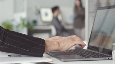 Ofis masasında dizüstü bilgisayarda yazan tanınmayan iş kadınının elleri. Ofis çalışanları arka planda sohbet ediyorlar.