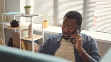 Genç siyahi adam modern ofiste masada oturuyor ve cep telefonuyla konuşuyor.