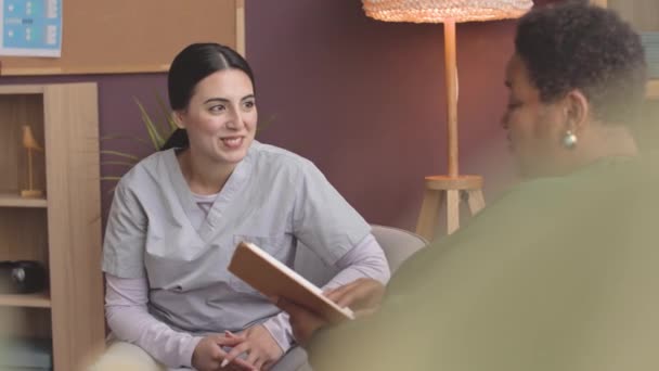 成熟的非洲裔美国妇女坐在疗养院的沙发上 手里拿着一本书与年轻的女护士交谈 — 图库视频影像