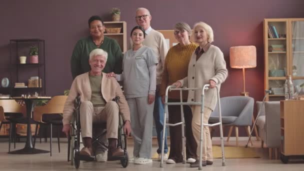 Stock video Locked down full length portrait of young female nurse in scrubs and ethnically diverse group of seniors posing for camera standing in resting room of cozy nursing home