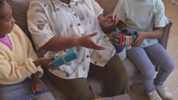 上記のショットから アフリカ系アメリカ人の子供2人の間に包まれたギフトボックスでプレゼントを贈り リビングルームでリビングルームで祖母の編み物をする — ストック動画