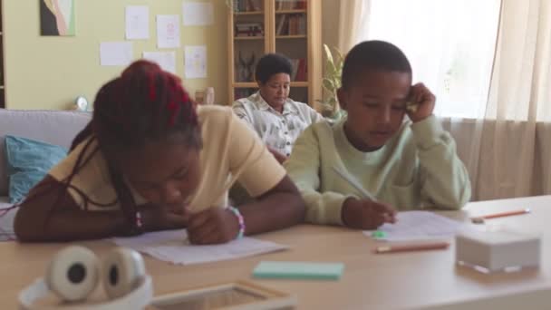 两个非裔美国人二年级的兄弟姐妹在客厅的书桌边做数学作业 而他们的祖母则坐在沙发上做背景介绍 — 图库视频影像