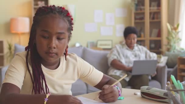 聪明的非洲裔美国女学生在客厅的课桌边做数学作业 而与奶奶一起呆在家里 用笔记本电脑坐在沙发上 背景模糊不清 — 图库视频影像