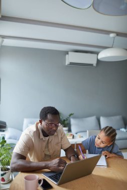 Evde çalışan Afro-Amerikan bir adamın dikey portresi ve yanında güzel bir kızı olan bir dizüstü bilgisayar kullanıyor.