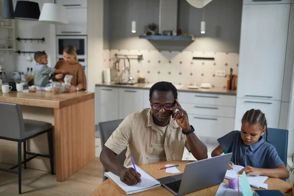 Front view of black father working from home with daughter sitting at table beside him, copy space