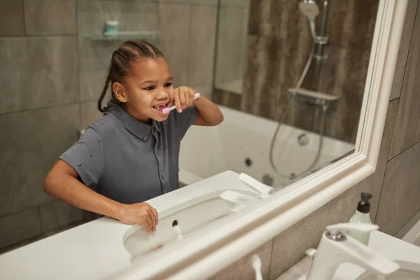 Mirror portrait of cute black girl brushing teeth with pink toothbrush good dental hygiene practice in childhood