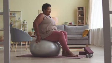 Artı, oturma odasında gümüş topun üzerinde otururken pembe spor kıyafetli yetişkin bir zenci kadın bacak egzersizi yapıyor.
