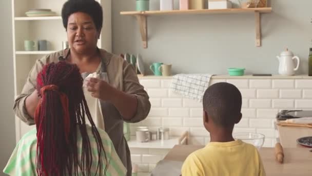 非洲裔美国人祖母给她的两个孙子女穿上围裙 准备在舒适的厨房里一起做饭 — 图库视频影像