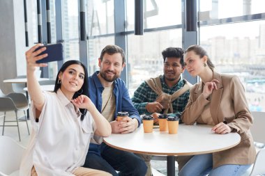 Bir grup yetişkin insan, kapalı kafede otururken selfie çekiyorlar.