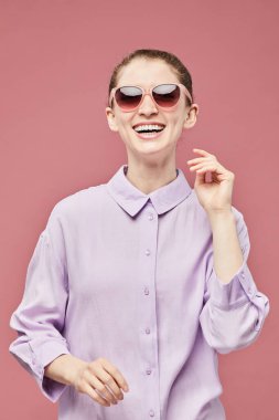 Güneş gözlüğü takan, pembe elbiseli gülümseyen genç bir kadının samimi portresi stüdyoda canlı pembe bir arka plana karşıydı.