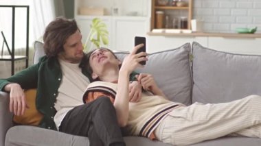 Romantik, genç, beyaz, eşcinsel çiftin, parlak, modern dairelerinde, yumuşak koltuklarda dinlenip, akıllı telefon ekranlarına bakmaları ve sohbet etmeleri.