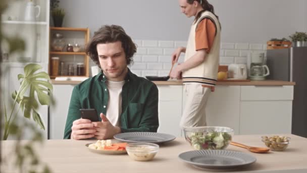 若いコーカサスゲイ男性はキッチンテーブルに座ってスマートフォンでブラウジングしながら 自宅で健康的なロマンチックな夕食を作ります — ストック動画