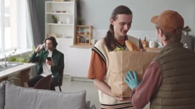 Modern Kafkasyalı eşcinsel çift evde yemek siparişi veriyor. Eldiven takan ve maske takan erkek kurye tarafından kağıt torbayla getiriliyor.