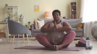 Orta yaşlı kıvrımlı siyah kadın nilüferde oturuyor yoga minderinde poz veriyor ve gözleri kapalı meditasyon yapıyor.