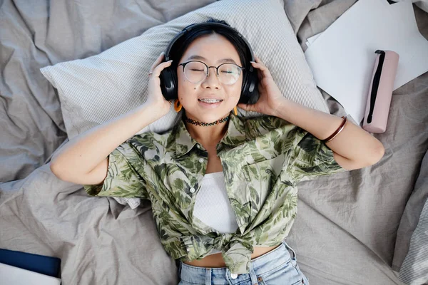 ヘッドフォンを着てベッドに横たわるアジアの十代の少女のトップビュー肖像画 勇敢なカメラに笑顔 — ストック写真