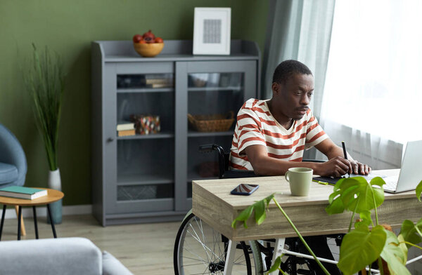 Portrait of adult black man in wheelchair using pen tablet while working with computer at home, copy space