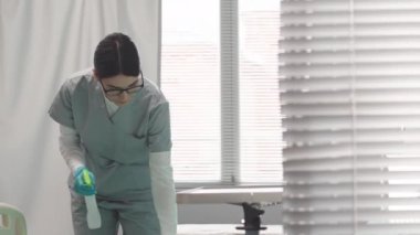 Genç Kafkas kadın hemşire hastane koğuşunu temizliyor, yatağı sterilize ediyor ve antiseptik ile diğer yüzeyleri temizliyor.