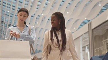 Modern alışveriş merkezinde ellerinde alışveriş torbalarıyla yürürken çok ırklı iki kadının sohbet ettiği orta boy bir fotoğraf.