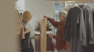İki çağdaş kadın lüks tasarımcı galerisinde bej örgülü elbiseyi askıda asılı tartışarak alışveriş yapıyorlar.