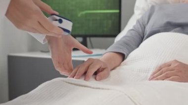 Tanınmayan hemşire oksijen doygunluğunu kontrol etmek için hastane yatağında yatan hastaya parmak nabız oksimetresi uyguluyor.