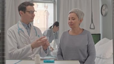 Genç erkek doktor, hastane odasında hemşire arka planda yatağı toplarken olgun Asyalı kadınlara hangi ilaçları almaları gerektiğini anlatıyor.