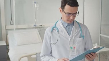 Beyaz laboratuvar önlüklü ve gözlüklü beyaz erkek doktorun hastane odasında elinde bir panoyla kameraya bakarken orta boy portresi.