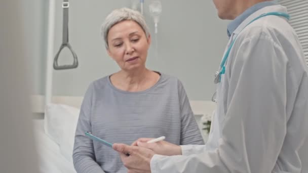 在医院病房与亚洲老年女性交谈时 将穿着白衣的年轻男性医生倾斜到病人医疗卡上做笔记 — 图库视频影像