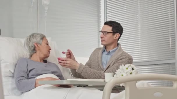 男人在医院病房照顾生病的母亲 用勺子喂她吃酸奶 — 图库视频影像