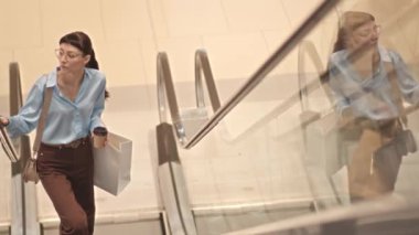 Alışveriş torbası ve kahve fincanı elinde genç beyaz kadın modern alışveriş merkezinde tek başına alışveriş yaparken yürüyen merdivenden yukarı çıkıyor.