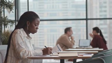 Panoramik pencereli bir kafede çalışırken finansal belgeleri analiz eden Afro-Amerikan genç bir kadının yan görüntüsü